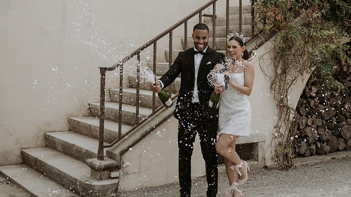 Engaged couple in wedding-inspired outfits popping a champagne bottle to celebrate their engagement